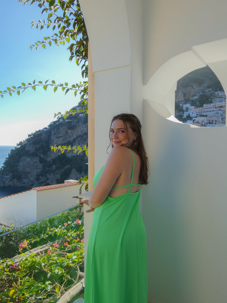 A photo of the founder wearing a bright green dress, with her back turned slightly to the camera, looking over her left shoulder and smiling softly. She is standing on a balcony outside, with a view of a coastal town in the background.